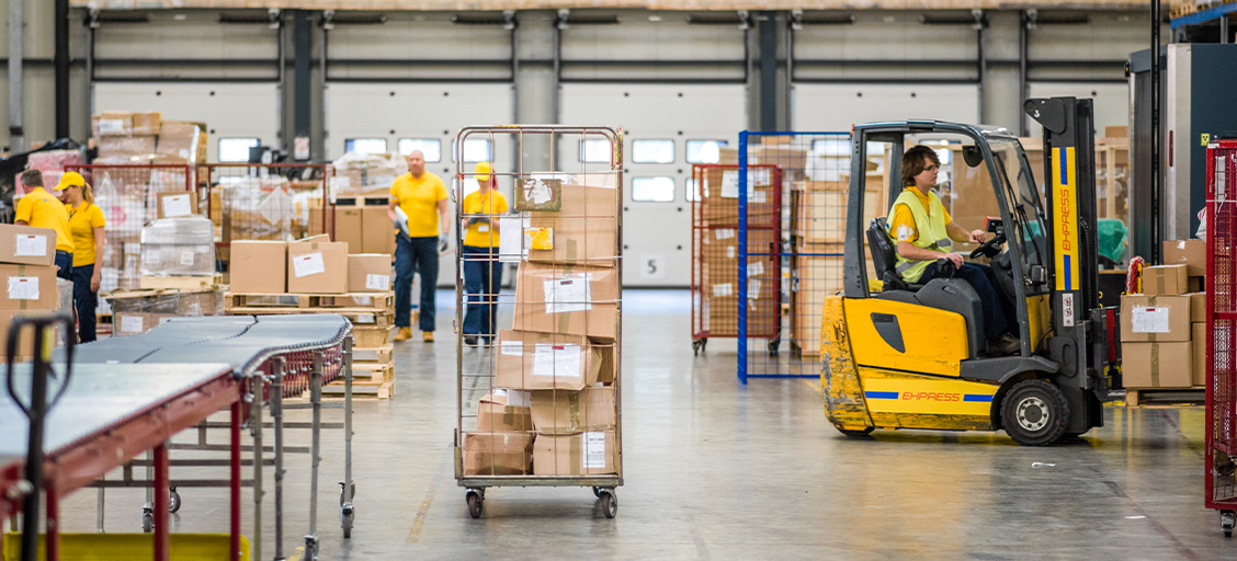 A person in a yellow safety vest driving a forklift stacked with boxes in a distribution center.