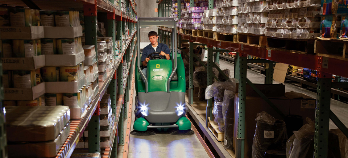 A person drives a green floor cleaning machine in between rows of shelving stacked with products in a warehouse.